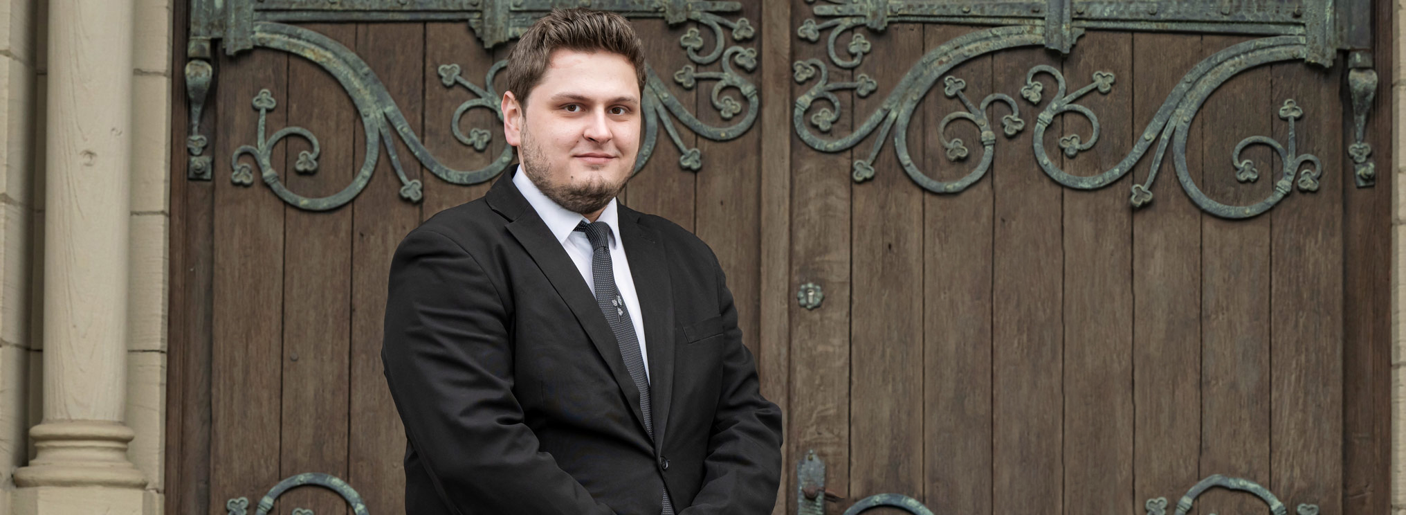Zwei Mitarbeiter von Gabriel Bestattungen aus dem Ruhrgebiet stehen vor einer kunstvoll verzierten Holztür. Beide tragen formelle, schwarze Anzüge und vermitteln Seriosität und Einfühlsamkeit. Das Bild symbolisiert die würdevolle Begleitung und Unterstützung in schweren Zeiten.